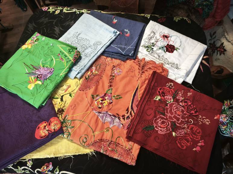 „Mehrere gefaltete Altardecken aus Leinen, handbestickt mit floralen Motiven, Tieren und ornamentalen Linien in verschiedenen Farben, ausgelegt auf einem dunklen Untergrund.“