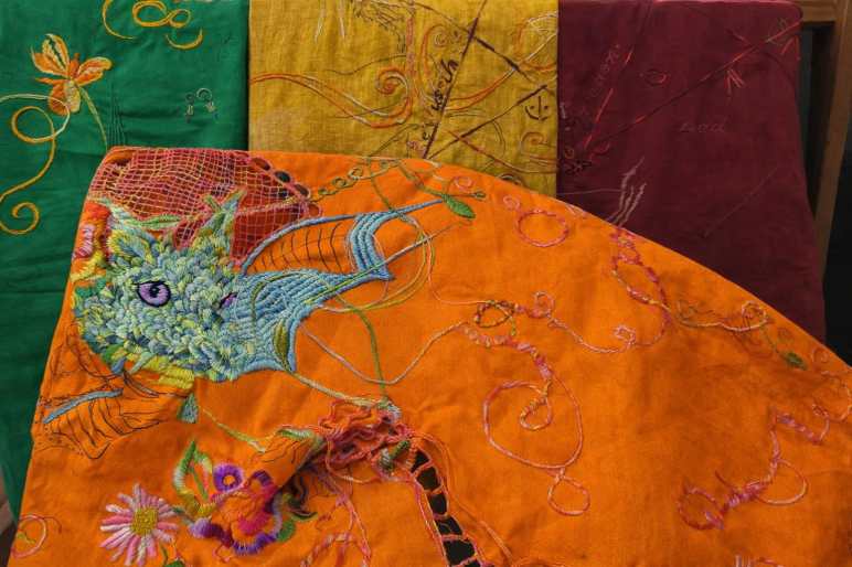 Handgestickter Drachenkopf auf orangefarbenem Stoff im Stickrahmen. Der Drache ist detailreich in Blau- und Grüntönen gearbeitet, mit violettem Auge. Um ihn herum florale Elemente und freie Fäden. Im Hintergrund liegen weitere farbige Stoffbahnen in Grün, Gelb und Rot.