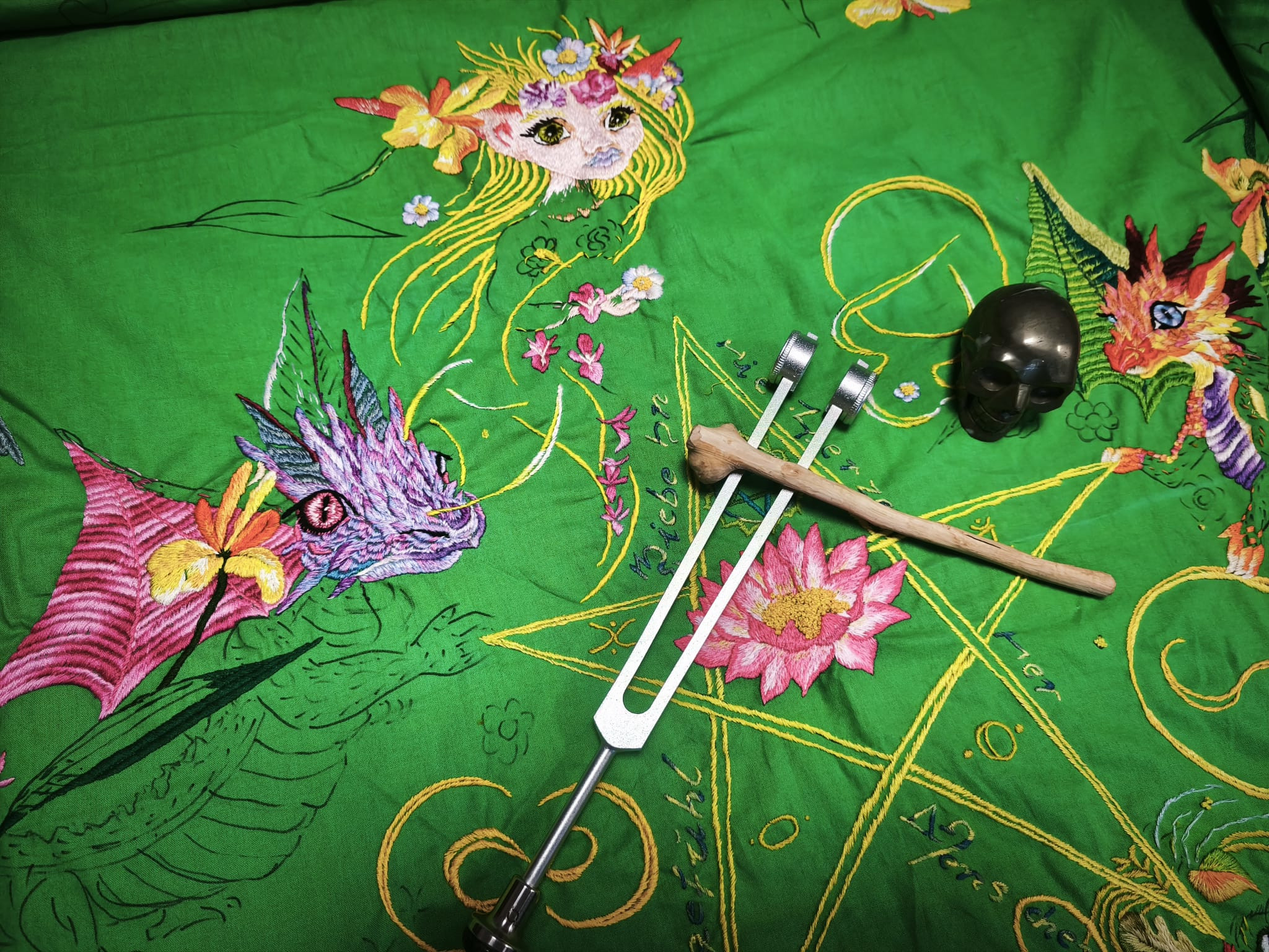 Grüne handbestickte Herzchakra-Decke in Arbeit mit Drachenmotiven, darauf eine Stimmgabel mit Holzschlägel und ein dunkler Heilsteinschädel, aus der praktischen Energie- und Körperarbeit heraus angeordnet