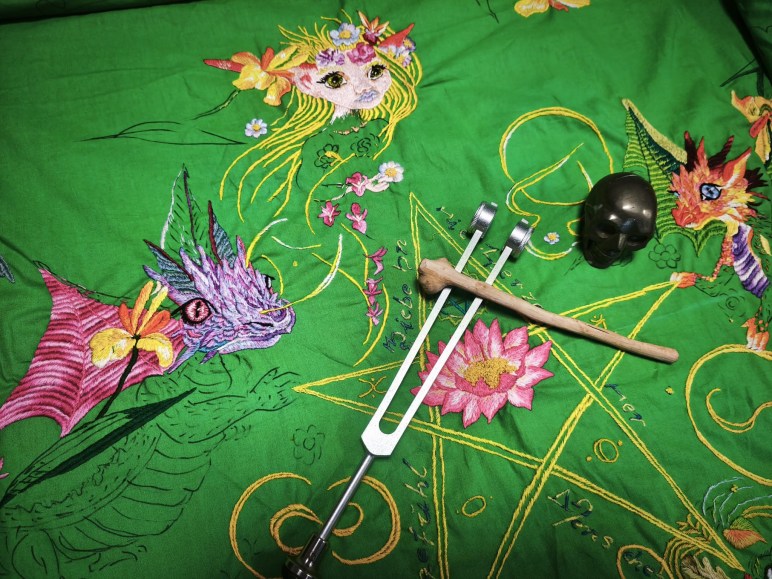 Grüne handbestickte Herzchakra-Decke in Arbeit mit Drachenmotiven, darauf eine Stimmgabel mit Holzschlägel und ein dunkler Heilsteinschädel, aus der praktischen Energie- und Körperarbeit heraus angeordnet